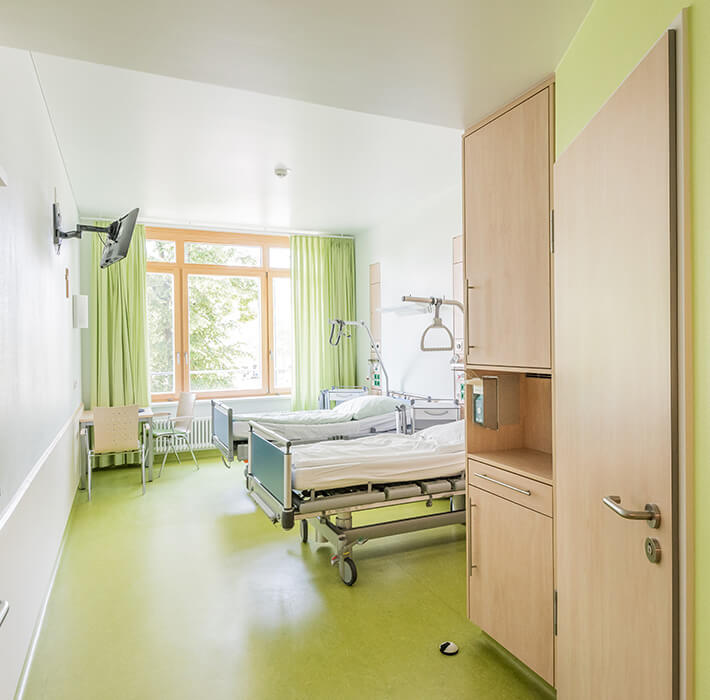 Krankenzimmer mit zwei Betten und einem Fernseher. Der Raum hat einen grünen Boden und grüne Vorhänge. Die Wände sind weiß und teilweise mit grün.