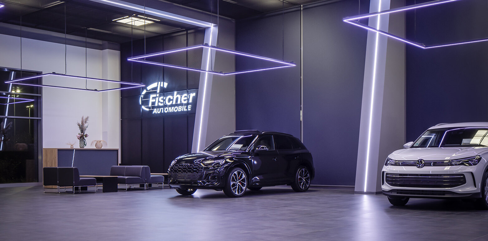 Mehrere Neuwagen von Volkswagen geparkt in moderner Fahrzeughalle mit eleganter Ausleuchtung anhand von quadratischer, linienförmiger Beleuchtung von oben. An der Wand in Leuchtschrift das Logo von Fischer Automobile