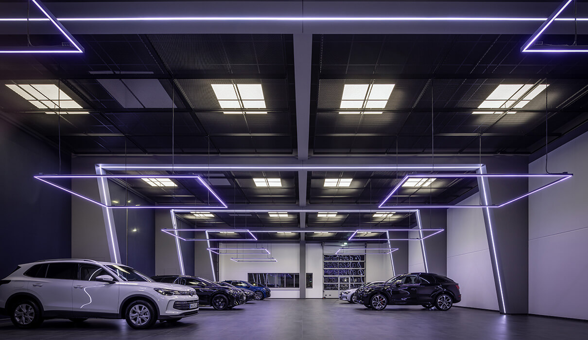 Mehrere Neuwagen von Volkswagen geparkt in moderner Fahrzeughalle mit eleganter Ausleuchtung anhand von quadratischer, linienförmiger Beleuchtung von oben.
