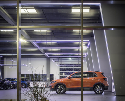Blick von außen in eine hochmoderne Fahrzeughalle. Ein orangefarbener Volkswagen ist von der Seite zu sehen.