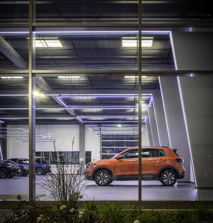 Blick von außen in eine hochmoderne Fahrzeughalle. Ein orangefarbener Volkswagen ist von der Seite zu sehen.