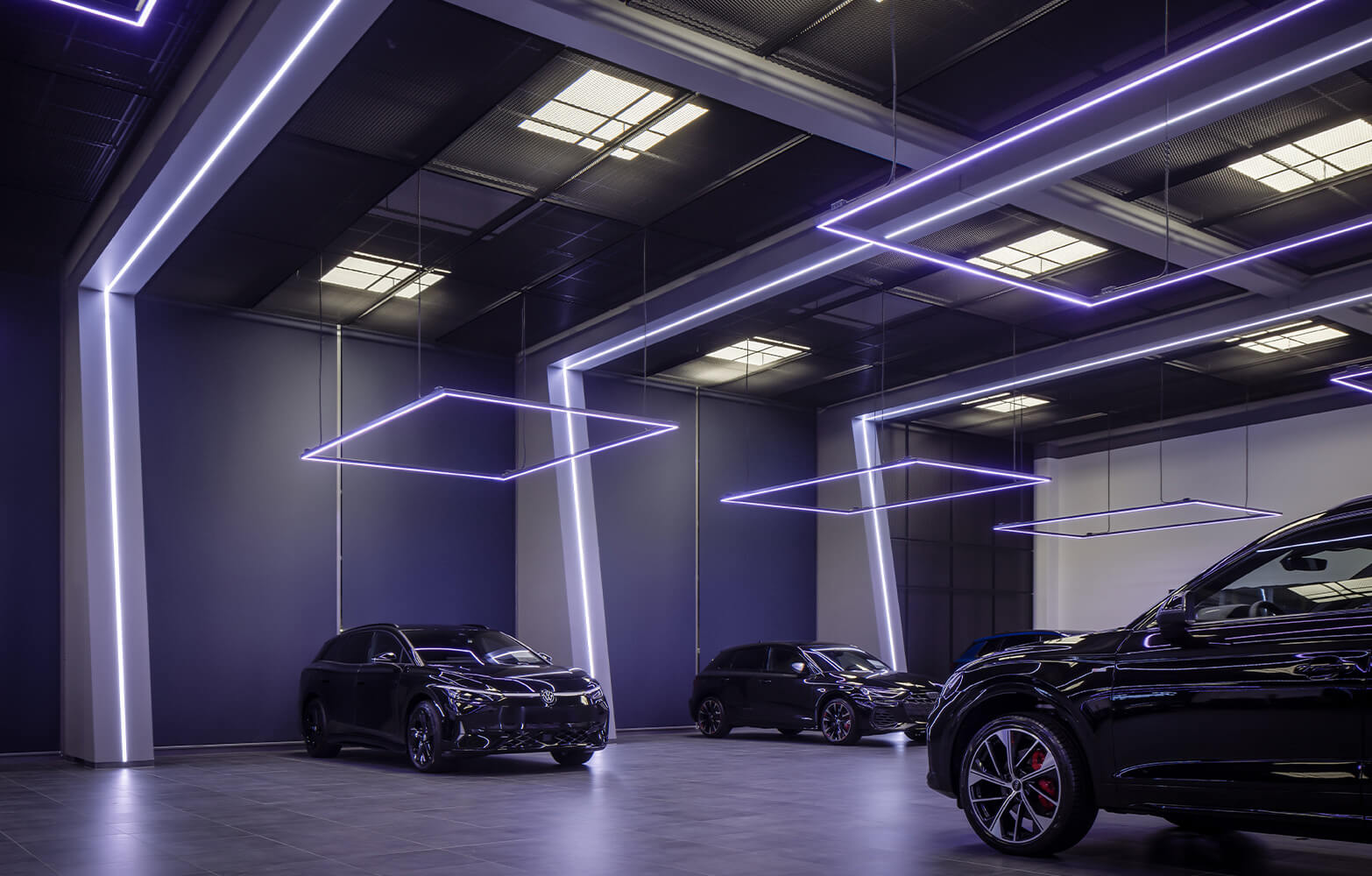 Mehrere schwarze Neuwagen von Volkswagen geparkt in moderner Fahrzeughalle mit eleganter Ausleuchtung anhand von quadratischer, linienförmiger Beleuchtung von oben.