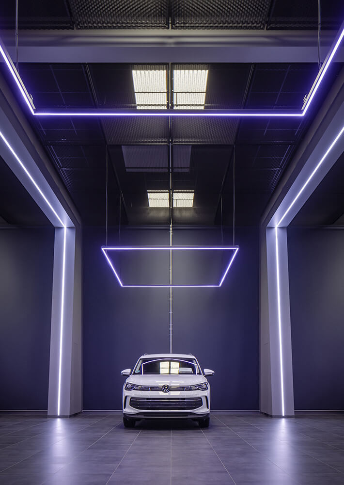 Weißes Auto von Volkswagen geparkt in moderner Fahrzeughalle mit eleganter Ausleuchtung anhand von quadratischer, linienförmiger Beleuchtung von oben.