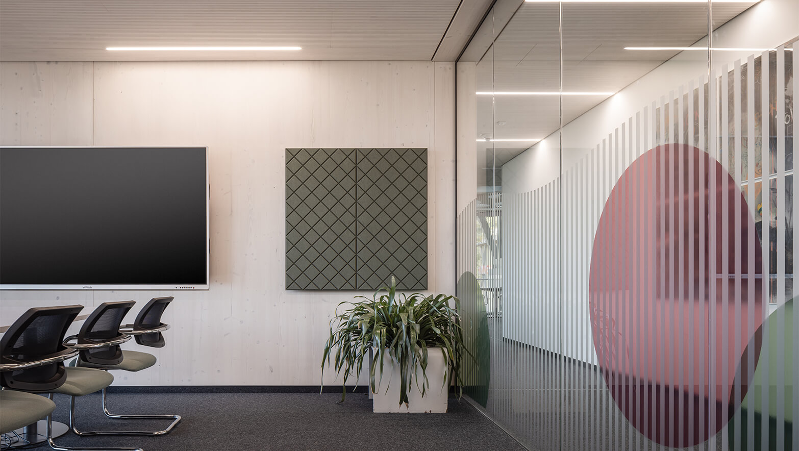 Blick in den Konferenzraum, link Stühle und Präsentationsmonitor, mitte eine Pflanze und ein Kunstwerk, rechts gestaltete Glaswand mit Streifen und Ornamenten