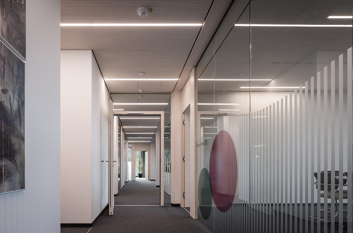 Blick in den Konferenzraum, link Stühle und Präsentationsmonitor, mitte eine Pflanze und ein Kunstwerk, rechts gestaltete Glaswand mit Streifen und Ornamenten