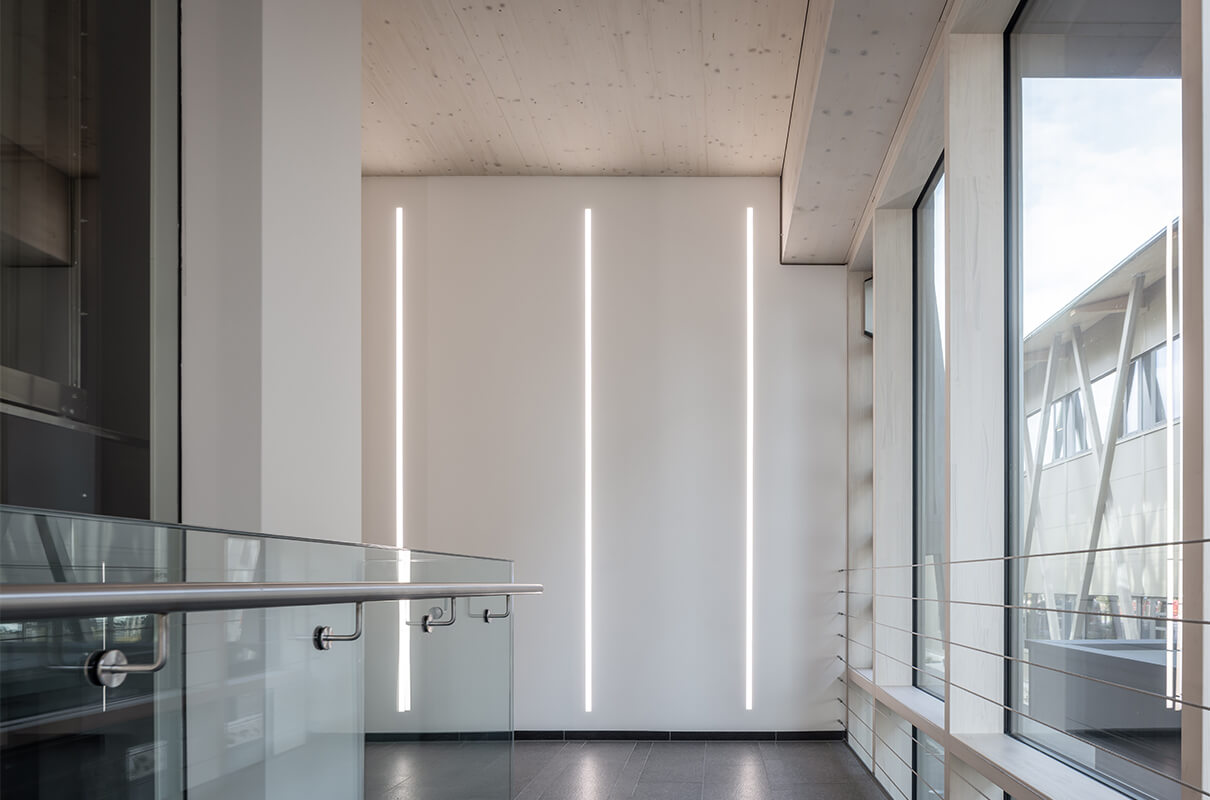 Helles Treppenhaus der Hammerbacher GmbH mit elegantem Lichtkonzept in Form von Lichtstreifen an den Wänden. Zu sehen sind hier auch der Handlauf, sowie ein Blick aus dem Fenster zum gegenüberliegenden Gebäude.